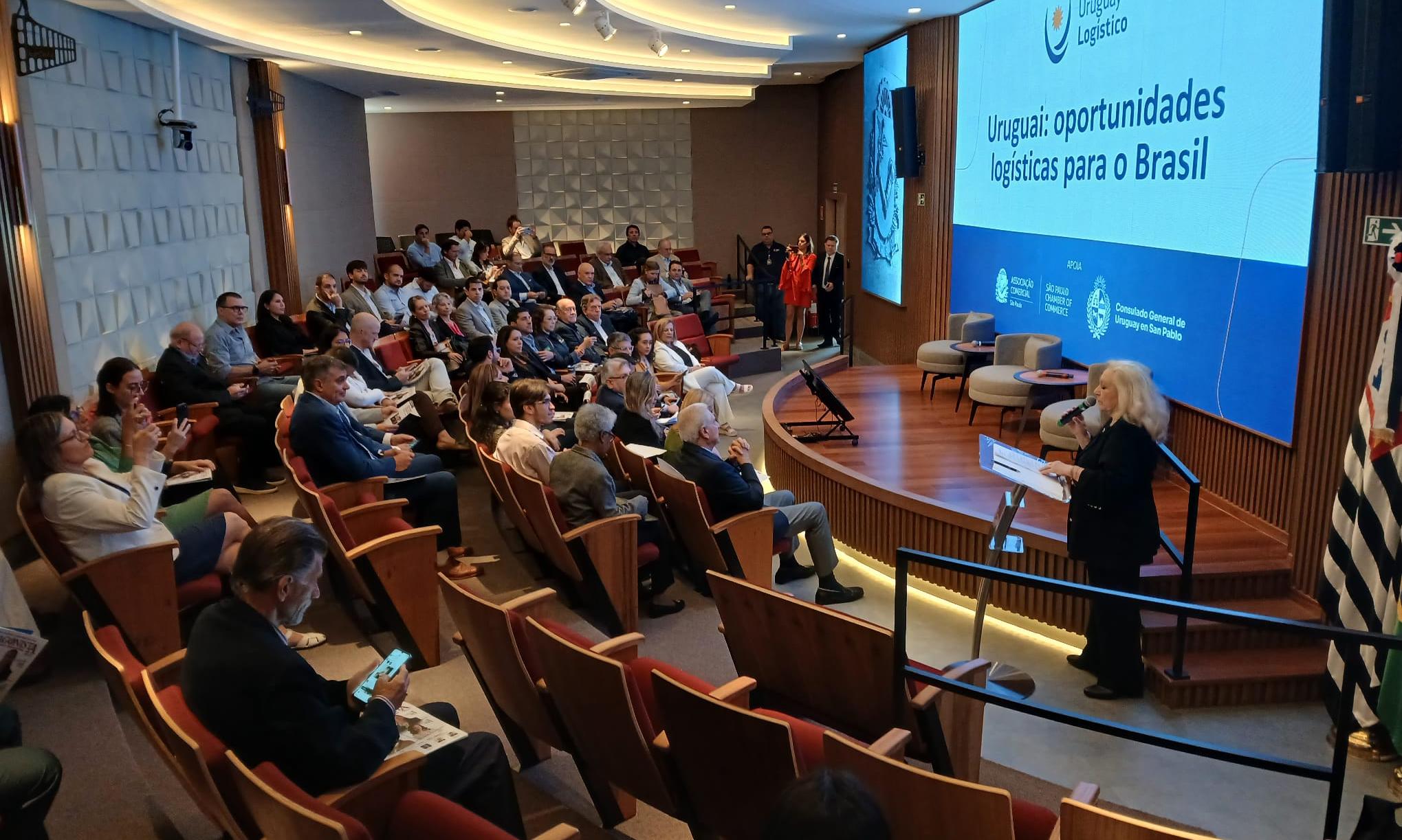 Auditorio en forma semicircular con personas sentadas y una gran pantalla en el escenario con el texto "Uruguai: oportunidades logísticas para o Brasil"